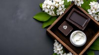Luxury spa setting with white cream in jar, dark wooden tray, spring blossom flowers and green leaves on black background. Wellness and skincare concept.