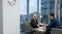 Two professionals engaged in business discussion in modern high-rise office with city skyline view. Caucasian woman and man in formal attire seated in contemporary lounge area, suitable for corporate, recruitment, and business communication concepts.