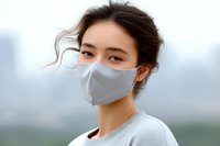 Young Asian woman wearing protective gray face mask outdoors with city skyline in background, wind blowing through dark hair, casual attire.