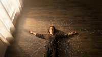 Young woman lying on wooden floor with arms spread wide surrounded by glowing light particles in warm sunlit room creating magical atmosphere.