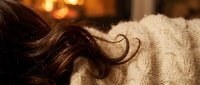 Young woman with long brown hair resting in cozy cream knit sweater by warm fireplace, capturing hygge winter comfort lifestyle.