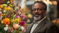 Mature African American businessman with glasses and beard smiling confidently in modern office with colorful floral arrangement, professional portrait.
