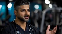 Middle Eastern male doctor in black scrubs with stethoscope gesturing while speaking, confident expression against blurred gym background with bokeh lights.