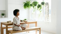 Young woman with curly dark hair and medium skin tone sitting on wooden bench at table, working on laptop in bright, minimalist workspace with hanging green plants and natural light.