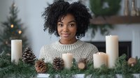 Young African American woman with natural curly hair smiling warmly while sitting at decorated Christmas table with white candles and pine cones, wearing cozy knit sweater.