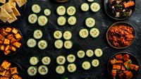 Fresh cucumber slices arranged in pattern on dark background with roasted sweet potato cubes and spicy sauce bowls, overhead view of healthy appetizer spread.