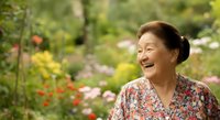 Middle-aged Asian woman smiling in blooming garden with colorful flowers, wearing floral dress, enjoying nature outdoors.