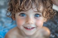 Close up portrait of a young child with bright blue eyes and natural curly brown hair smiling joyfully against soft blurred background. Genuine happiness expression.