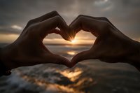 Hands forming heart shape silhouette against golden sunset over ocean water creating romantic love symbol for Valentine's Day greeting cards.