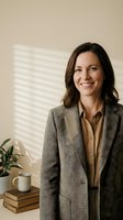 Professional businesswoman in gray blazer smiling confidently in modern office setting with natural lighting and minimalist decor.