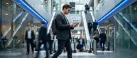 Professional businessman in dark suit using smartphone while walking through modern office building lobby with glass architecture and blurred people in background.