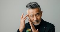Mature man with gray hair and facial hair wearing black jacket, hand to face in thoughtful pose against neutral background.