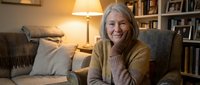 Senior woman with long gray hair and Caucasian ethnicity sitting in comfortable armchair in cozy home library, wearing mustard cardigan, smiling at camera with warm lighting and bookshelves.