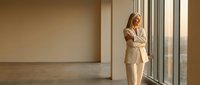 Professional blonde businesswoman in beige suit standing by office windows with crossed arms, confident corporate executive in modern workplace interior.