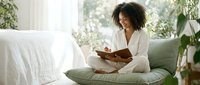 Young African American woman reading book while sitting on bed in bright bedroom with plants, relaxing morning routine and self-care lifestyle.
