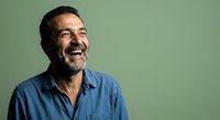 Middle-aged man with dark hair and beard wearing light blue shirt, smiling and looking away against green background for portrait and lifestyle use.