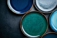 Handcrafted ceramic plates in vibrant blue and turquoise glazes arranged on dark stone background, showcasing artisanal pottery design and modern tableware styling.