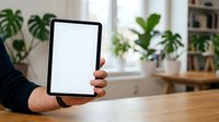 Person holding tablet with blank screen in modern office workspace surrounded by potted plants and wooden furniture for app mockup and tech presentation.
