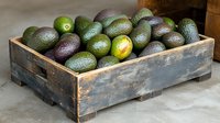Fresh ripe avocados in rustic wooden crate on concrete surface, mix of green and dark purple fruits showing natural ripening stages for food market.