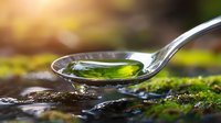 Shiny metal spoon with green liquid medicine or essential oil drop against natural moss background, macro photography with water drops and sunlight.