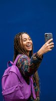 Happy young African American woman with braids taking selfie with smartphone wearing purple backpack against blue background for social media content.