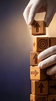 Hands stacking wooden blocks with business symbols including arrows, target, and house icons representing growth strategy and goal achievement concepts.