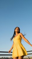 Young woman with long dark braided hair and medium brown skin tone wearing bright yellow sleeveless dress standing outdoors against clear blue sky, lifestyle and fashion photography.