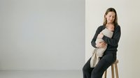 Young mother in dark casual wear holding sleeping newborn baby while sitting on wooden stool against neutral beige background in minimalist studio setting.
