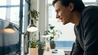 Happy Caucasian businessman smiling while working on computer in modern office with plants. Professional male entrepreneur focused on screen in workplace.