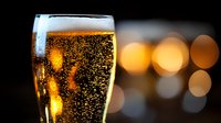 Golden bubbling beer in pint glass with rising carbonation against blurred bokeh lights background, refreshing alcoholic beverage close-up photography.