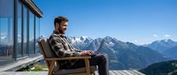 Young businessman working remotely on laptop from mountain terrace with alpine peaks backdrop, enjoying digital nomad lifestyle and scenic workspace.