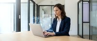 Professional businesswoman working on laptop in modern office environment with natural lighting and glass partitions for corporate presentations.