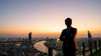 Professional businessman silhouette standing on rooftop overlooking illuminated city skyline at sunset with river view for corporate success concepts.