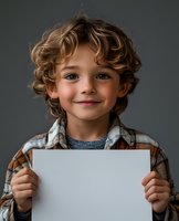 Young child with curly blonde hair wearing plaid shirt holding blank white paper against dark background, gentle smile and direct gaze at camera.