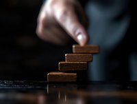 Hand stacking wooden blocks in ascending order against dark background, creating steps or stairs metaphor for growth, progress and business success.