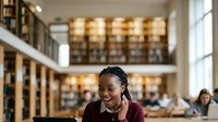 Young woman with dark skin tone and braided hairstyle wearing burgundy sweater and white collar, smiling while studying in modern library with bookshelves and natural light from windows.
