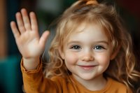 Cheerful young child with blonde curly hair wearing orange sweater waves hello with bright smile and joyful expression in natural indoor lighting.