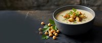 Creamy chickpea soup with herbs and spices in ceramic bowl on dark slate surface, healthy vegetarian meal with scattered legumes and fresh parsley garnish.