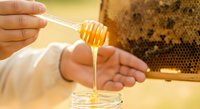 Beekeeper in white protective suit holding honey dipper with golden honey dripping into jar, honeycomb frame visible in background, raw honey production and beekeeping concept.