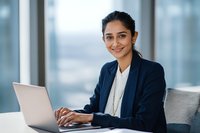 Confident South Asian businesswoman in navy blazer working on laptop in modern office with large windows, representing corporate success.