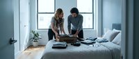 Young couple packing luggage on bed in modern bedroom with large window, preparing for travel or relocation.