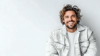 Smiling man with curly dark hair wearing white denim jacket and t-shirt against light background. Isolated portrait showing genuine happiness and confidence.