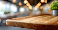 Wooden cutting board corner on blurred restaurant background with warm bokeh lights and decorative plant, close up view of natural wood grain texture.