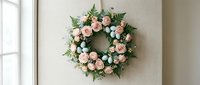 Elegant floral wreath with soft pink roses and eucalyptus leaves hanging on neutral wall near window for home decor and seasonal decoration.