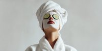 Young woman in white bathrobe and towel turban relaxing with cucumber slices on eyes during spa treatment, self-care routine on neutral background.