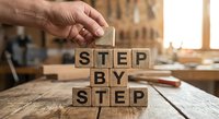 Hand placing wooden letter blocks spelling STEP BY STEP on rustic table in workshop setting for business planning and goal achievement concepts.