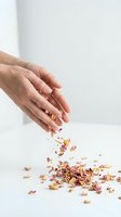 Female hand with gold and pink nail art sprinkling dried flower petals onto white surface for wellness, skincare, and natural beauty product marketing.