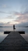 Silhouette of person practicing yoga tree pose on wooden dock at misty lake during peaceful sunrise with dramatic cloudy sky for wellness and meditation.