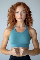 Young athletic woman with natural red curly hair wearing turquoise sports bra poses confidently against light background, showcasing fitness and wellness lifestyle.