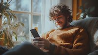Young Caucasian man with curly hair wearing orange sweater smiling while using smartphone, sitting on couch near window. Cozy home atmosphere with natural lighting.